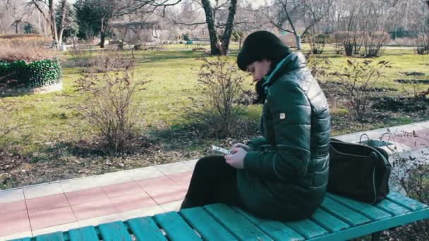 Дівчина друкує повідомлення і прокручує сторінки на смартфоні у весняному парку — стокове відео