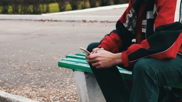 Solitario en una chaqueta roja está sentado en el parque y escribir algo en un teléfono inteligente — Vídeo de stock
