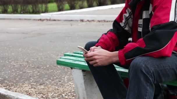 Un hombre solitario se sienta en un parque en un banco escribiendo algo en el teléfono inteligente — Vídeo de stock