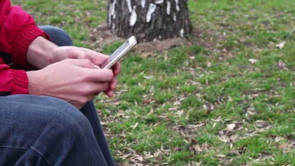 Young man with white phone sits in on street, in park, outdoor — Stock Video