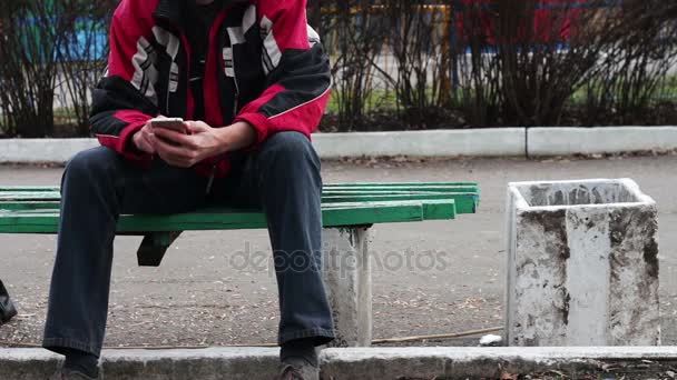 Un hombre solitario se sienta en un parque en un banco escribiendo algo en el teléfono inteligente — Vídeos de Stock