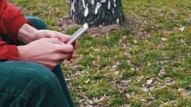 Lonely in a red jacket is sitting in the park and typing something on a smartphone — Stock Video