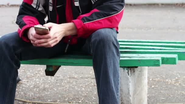 Un hombre solitario se sienta en un parque en un banco escribiendo algo en el teléfono inteligente — Vídeos de Stock