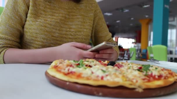 Mädchen in gelbem Pullover sitzt in einem Café und tippt etwas auf ein Smartphone, bevor auf dem Tisch eine leckere frisch zubereitete italienische Pizza liegt — Stockvideo