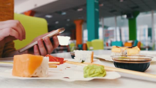 Meisje zit te typen op een smartphone en communiceren met de gesprekspartner in het café op de tafel zijn sushi, Japanse keuken — Stockvideo