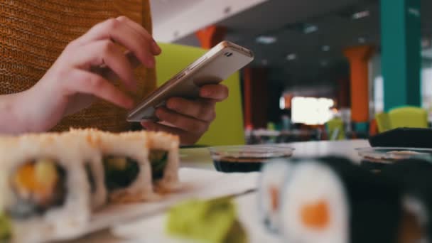 Menina senta-se digitando em um smartphone e se comunicar com o interlocutor no café na mesa são sushi, cozinha japonesa — Vídeo de Stock