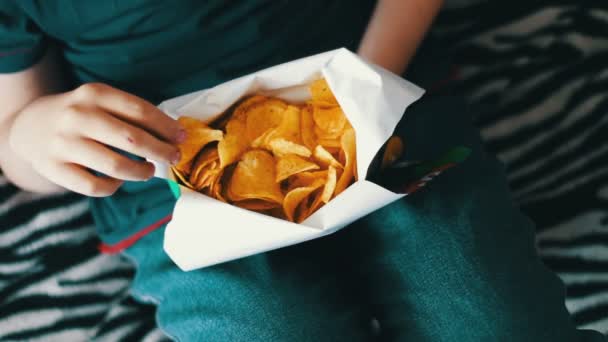 Menino adolescente comendo batatas fritas com as mãos no sofá em casa — Vídeo de Stock