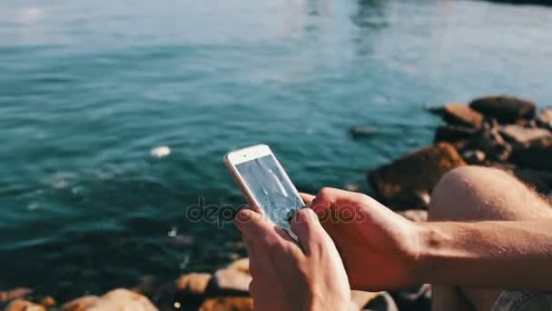 Kezében egy fiatalember írja be valamit egy okostelefon, a háttérben a Kaszpi-tenger hullámai — Stock videók