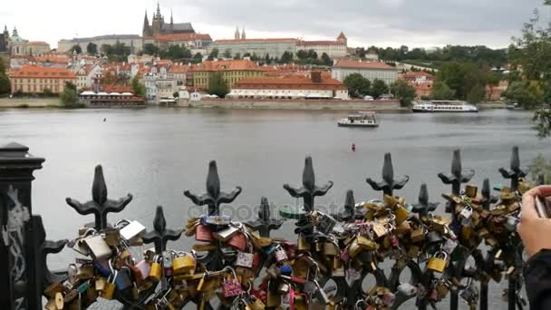 Vista da cidade velha de Praga e do rio Vltava, e Barcos passar, uma menina de pé na ponte com um monte de fechaduras de ferro e fotografias na paisagem magnífica smatrfon — Vídeo de Stock