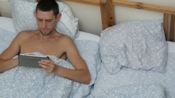 若い男はレプトンやストレッチング、あくびベッドに横たわってタブレットに何かを入力します。 — ストック動画