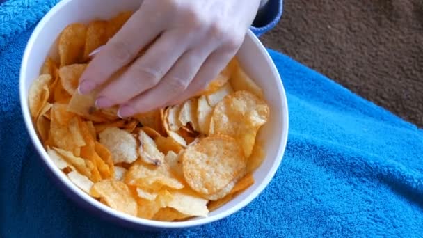 A mulher está em um sofá sob um cobertor azul e come batatas fritas de um prato profundo. Comida não saudável, fast food — Vídeo de Stock