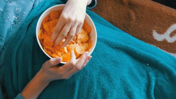 Donna è sdraiato su un divano sotto una coperta blu e sta mangiando patatine fritte — Video Stock