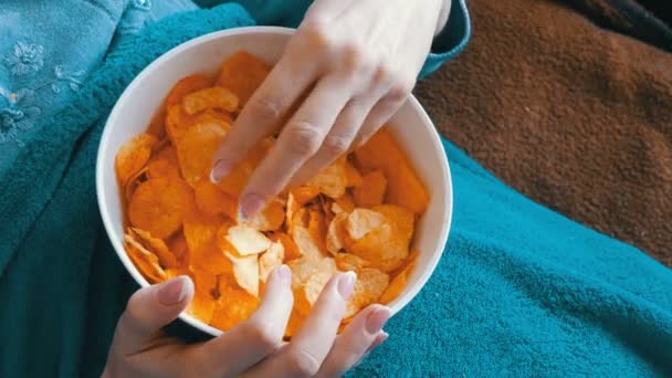 Mujer está acostada en un sofá debajo de una manta azul y está comiendo patatas fritas — Vídeos de Stock