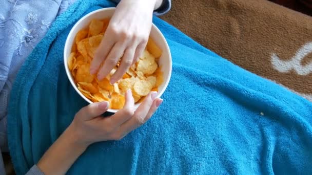 Kvinnan ligger på en soffa under en blå filt och äter potatis chips från en djup tallrik. Ohälsosam mat, snabbmat — Stockvideo