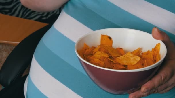 Um homem gordo e preguiçoso está a comer batatas fritas numa cadeira. Uma barriga enorme de um homem debaixo de uma t-shirt listrada. Fast food, não comida saudável — Vídeo de Stock