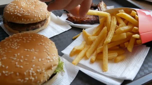 As mãos de um adolescente tomam batatas fritas. Hambúrgueres, asas de frango, batatas fritas na bandeja num restaurante de fast food. Alimentos não saudáveis — Vídeo de Stock