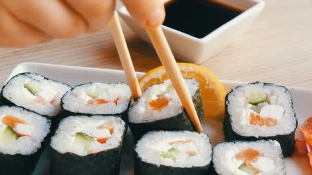 Un adolescente toma un rollo de sushi con palos de bambú chinos y lo deja caer en salsa de soja, toma un trozo de jengibre rosa. Cocina japonesa en plato de porcelana blanca junto al jengibre wasabi verde y salsa — Vídeos de Stock