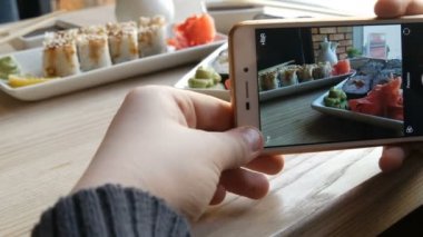 Eller genç bir Smartphone'da yiyecek bir fotoğraf olun. Bir dizi suşi rulo Japon mutfağı porselen çaydanlık soya sosu ve fincan tabağı için arka plan üzerinde şık bir kafede