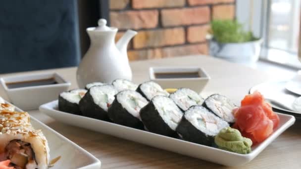 Два набора суши-роллов в стильном, декорированном цветами кафе. Ресторан японской кухни на столе рядом с суши фарфоровый чайник для соевого соуса и блюдца — стоковое видео
