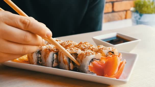 En tonåring pojke tar sushi rulle med kinesisk Bambu pinnar och droppar det in sojasås, tar en bit rosa ingefära. Japanska köket på vitt porslin tallrik bredvid gröna wasabi ingefära och sås — Stockvideo