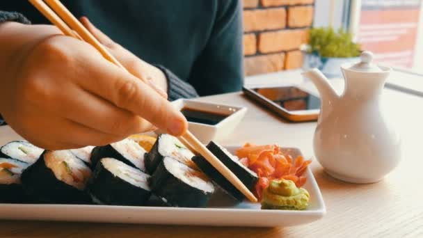 Een tiener jongen neemt sushi roll met Chinese bamboe stokken en het druppels in sojasaus, neemt een stuk Roze gember. Japanse keuken op witte porseleinen plaat naast groene wasabi, gember en saus — Stockvideo