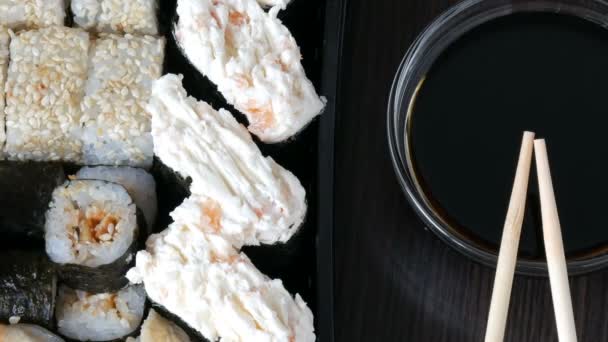 Стильно укладываемые суши на черном деревянном фоне рядом с соевым соусом и китайскими бамбуковыми палочками. Различные суши с лососем, угрем, огуречными креветками и другими разнообразными начинками — стоковое видео