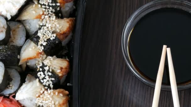Kamera rückt an. Stilvoll verlegtes Sushi auf schwarzem Holzgrund neben Sojasauce und chinesischen Bambusstäben. verschiedene Sushi-Rollen mit Lachs, Aal, Gurkengarnelen und anderen verschiedenen Füllungen — Stockvideo