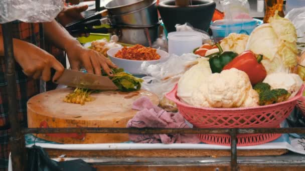 Woman cuts greens on a kitchen board with a large knife. Next to vegetables and cooking utensils. Thai street food — Stock Video