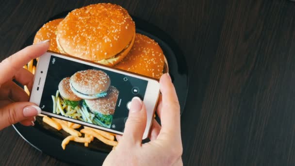 Las manos femeninas hacen la foto de un smartphone de una hamburguesa grande en un plato. Foto de comida rápida — Vídeos de Stock