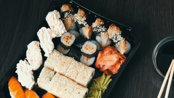 Elegantemente establecido sushi conjunto sobre un fondo de madera negro junto a la salsa de soja y palos de bambú chino. Varios rollos de sushi con salmón, anguila, camarones de pepino y otros rellenos diversos — Vídeos de Stock