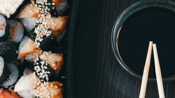 Camera moves up. Stylishly laid sushi set on a black wooden background next to soy sauce and Chinese bamboo sticks. Various sushi rolls with salmon, eel, cucumber shrimps and other various stuffing — Stock Video