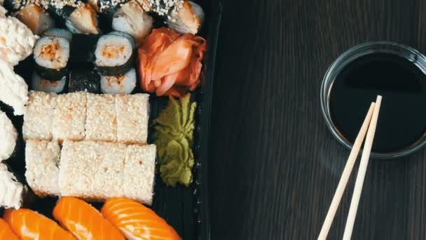 Des sushis élégamment posés sur un fond en bois noir à côté de la sauce soja et des bâtonnets de bambou chinois. Divers rouleaux de sushi au saumon, anguille, crevettes concombre et autres farces diverses — Video
