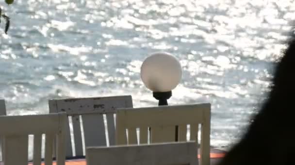 Έρημη καφέ με άσπρο ξύλινες καρέκλες στην παραλία με φόντο κύματα που χύνεται στον ήλιο. Πίνακας Cafe στην παραλία — Αρχείο Βίντεο