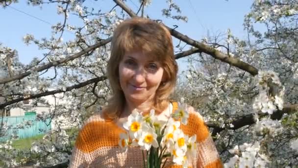 Día de las madres. Retrato de una hermosa mujer de mediana edad de ojos azules que felizmente mirando a la cámara, sonriendo, respira fragancia de flores en el fondo de un árbol floreciente en la primavera — Vídeos de Stock