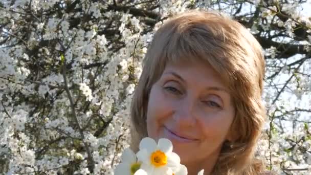 Ritratto di una bella donna di mezza età dagli occhi azzurri che guarda felicemente la macchina fotografica, sorridente, respira profumo di fiori sullo sfondo di un albero rigogliosamente fiorito in primavera. Festa della mamma — Video Stock