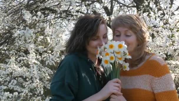Sehr schöne Mutter Alter und erwachsene Tochter mit einem Blumenstrauß umarmen lächelnd lächelt vor dem Hintergrund eines blühenden Baumes in die Kamera — Stockvideo