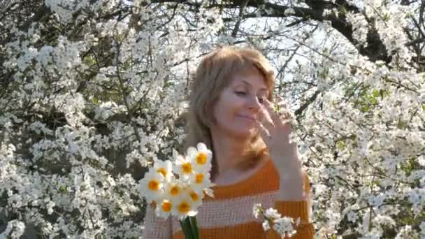 Портрет красивої блакитноокої жінки середнього віку, яка щасливо дивиться на камеру, усміхаючись, дихає ароматом квітів на тлі соковито квітучого дерева навесні. День матері — стокове відео