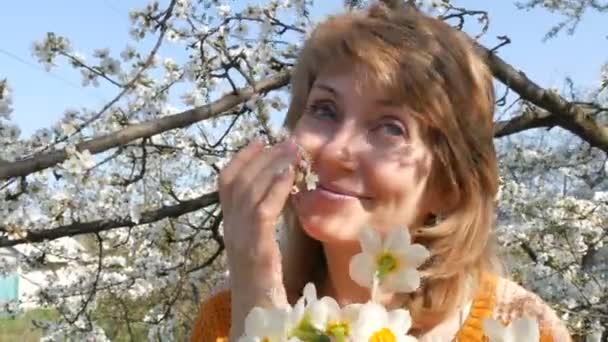 Festa della mamma. Ritratto di una bella donna di mezza età dagli occhi azzurri che guarda felicemente la macchina fotografica, sorridente, respira profumo di fiori sullo sfondo di un albero rigogliosamente fiorito in primavera — Video Stock