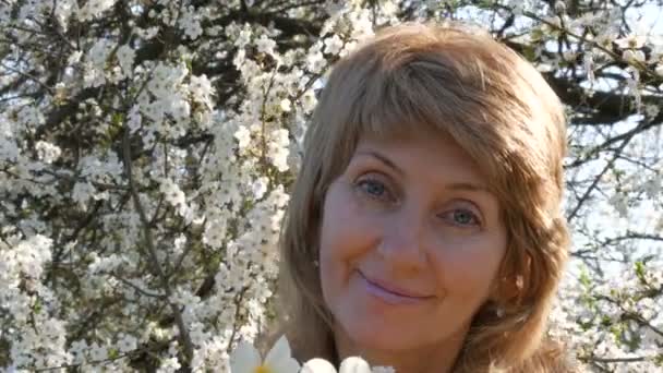 Retrato de una hermosa mujer de mediana edad de ojos azules que felizmente mirando a la cámara, sonriendo, respira fragancia de flores en el fondo de un árbol floreciente exuberante en la primavera. Día de las madres — Vídeos de Stock