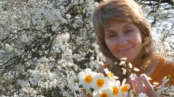 Портрет красивої блакитноокої жінки середнього віку, яка щасливо дивиться на камеру, усміхаючись, дихає ароматом квітів на тлі соковито квітучого дерева навесні. День матері — стокове відео