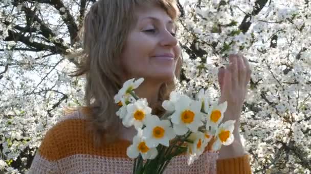 Retrato de una hermosa mujer de mediana edad de ojos azules que felizmente mirando a la cámara, sonriendo, respira fragancia de flores en el fondo de un árbol floreciente exuberante en la primavera. Día de las madres — Vídeos de Stock