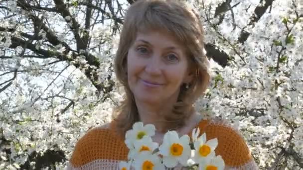 Hermosa mujer en años sosteniendo un ramo de narcisos mira a la cámara y sonríe. Una mujer con bellos, inusuales, bondadosos ojos azules está de pie sobre el fondo de un árbol en flor. Día de las Madres — Vídeos de Stock