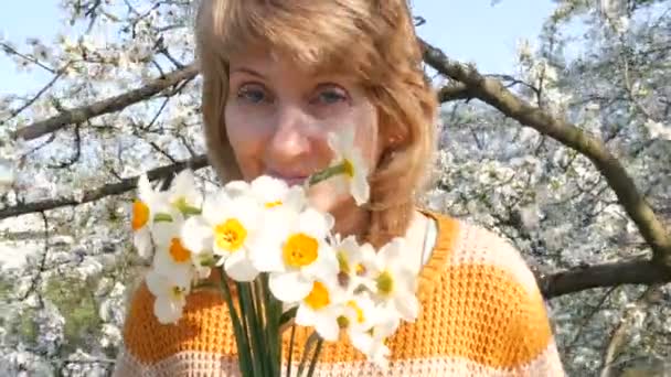 Día de las madres. Retrato de una hermosa mujer de mediana edad de ojos azules que felizmente mirando a la cámara, sonriendo, respira fragancia de flores en el fondo de un árbol floreciente en la primavera — Vídeos de Stock