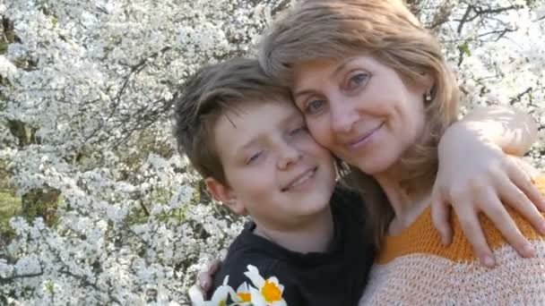 一个成年的中年母亲拥抱和亲吻她的十几岁的儿子温柔地在一个美丽的茂密的树的背景。春季母亲节 — 图库视频影像