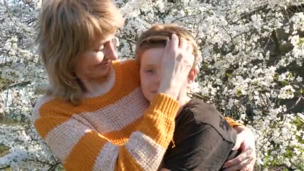 Взрослая мать среднего возраста обнимает и нежно целует своего сына-подростка на фоне красивого роскошного дерева. День матери весной — стоковое видео