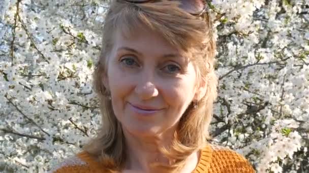 Retrato de una hermosa mujer de mediana edad de ojos azules que felizmente mirando a la cámara, sonriendo, respira fragancia de flores en el fondo de un árbol floreciente exuberante en la primavera. Día de las madres — Vídeos de Stock