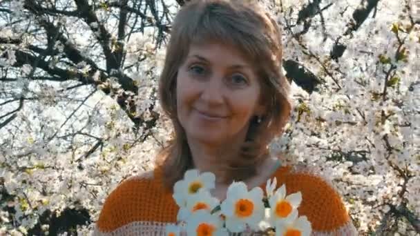 Día de las madres. Retrato de una hermosa mujer de mediana edad de ojos azules que felizmente mirando a la cámara, sonriendo, respira fragancia de flores en el fondo de un árbol floreciente en la primavera — Vídeos de Stock