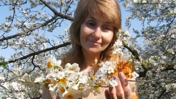 Festa della mamma. Ritratto di una bella donna di mezza età dagli occhi azzurri che guarda felicemente la macchina fotografica, sorridente, respira profumo di fiori sullo sfondo di un albero rigogliosamente fiorito in primavera — Video Stock