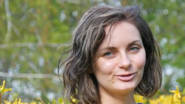 Beautiful happy girl with short hair looks at camera and smiles against the background of spring crowded bushes — Stock Video