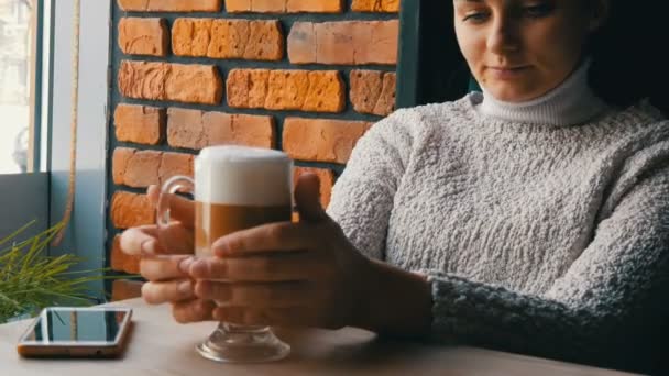 Güzel kız ikindilerde içecekler kahve latte hava köpük ile gülümseyen rahat kafede oturur ve pencereden görünüyor — Stok video